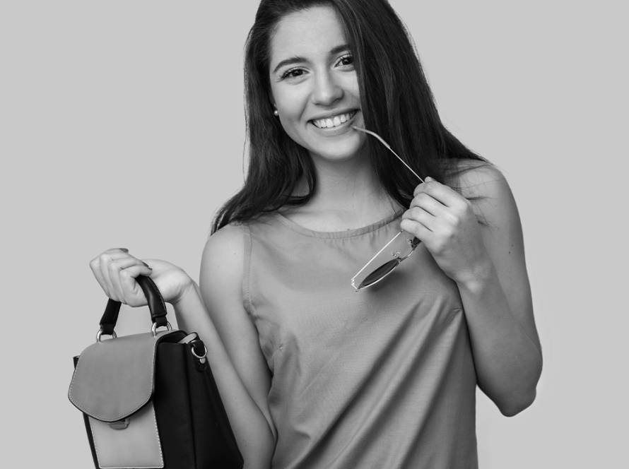 Joven sonriente con pelo largo y oscuro, sosteniendo un moderno y elegante bolso negro y gris y unas gafas de sol, sobre un fondo gris neutro, representando un estilo de vida despreocupado y los viajes con Lock & Relax, la empresa líder en consigna de equipajes en Málaga.