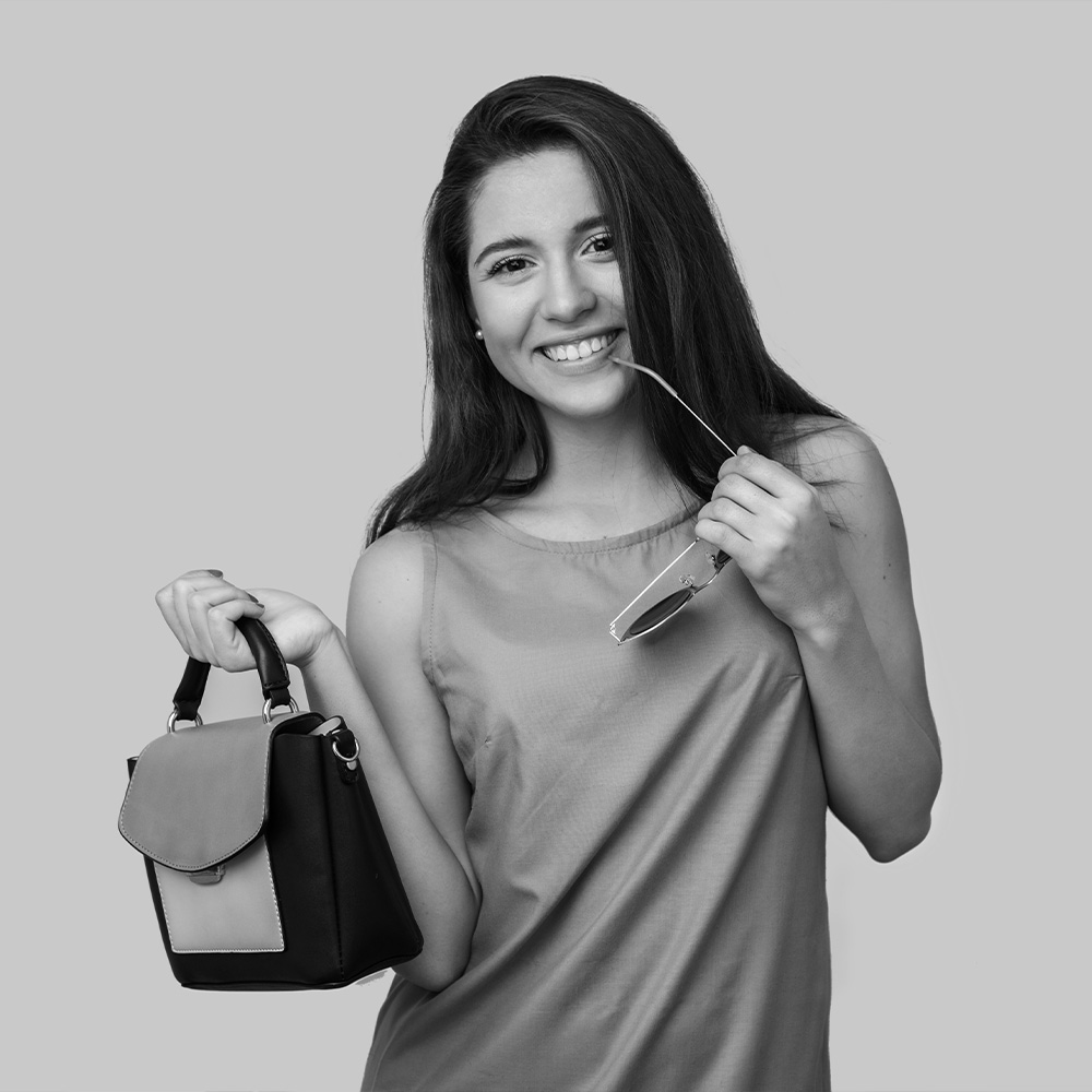 Smiling young woman with long dark hair, holding a stylish black and gray modern handbag and sunglasses, against a neutral gray background, representing a carefree lifestyle and travel with Lock & Relax, the leading luggage storage company in Málaga.